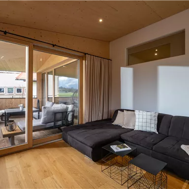 Ein modernes Wohnzimmer mit einer bequemen Couch und großen Fenstern. Draußen ist eine Terrasse mit einem Schaukelstuhl und einer gemütlichen Sitzgelegenheit.