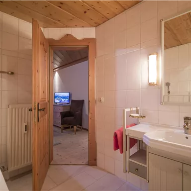 A modern bathroom with bright tiles and a sink. Through the open door, a cozy living area with a television is visible.