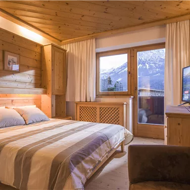 A cozy bedroom with wooden paneled walls and a large window that offers a view of the mountains. The room is modern and invitingly furnished, with a television and a comfortable armchair.