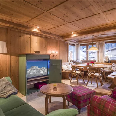A cozy living room with wooden paneling and a large dining table. There is a green couch, a round table, and lots of natural light coming through the windows.