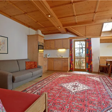 Ein helles Wohnzimmer mit Holzdecke und einem roten Teppich. Es gibt eine Couch, eine Küchenzeile und einen Essbereich mit Fenster.