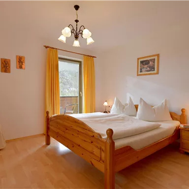A cozy bedroom with a wooden bed and two pillows. The room has large windows with yellow curtains and warm light.