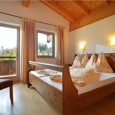 Ein gemütliches Schlafzimmer mit einem Doppelbett und Holzmöbeln. Große Fenster lassen viel Tageslicht herein und bieten einen schönen Blick nach draußen.