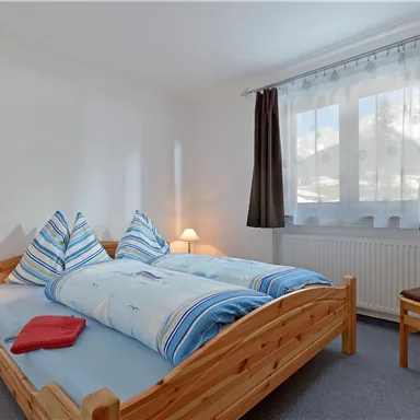A cozy bedroom with a double bed and two nightstands. The room has light-colored walls and a window with curtains.