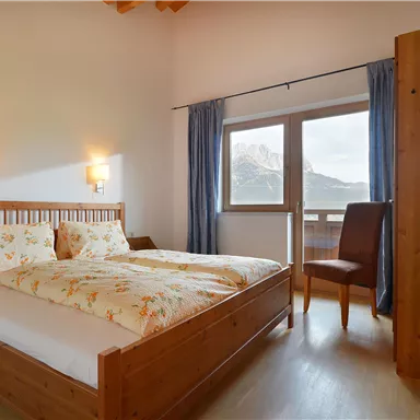 Ein helles Schlafzimmer mit einem Doppelbett und floralen Bettwäsche. Vor dem Fenster eröffnet sich ein Blick auf die Berge.