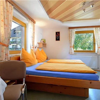 A cozy bedroom with two beds and orange pillows. The room has large windows that let in a lot of daylight.