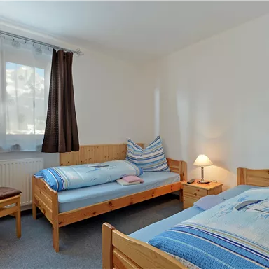 A simple bedroom with two single beds and a chair. There is a window with curtains and a bedside lamp.