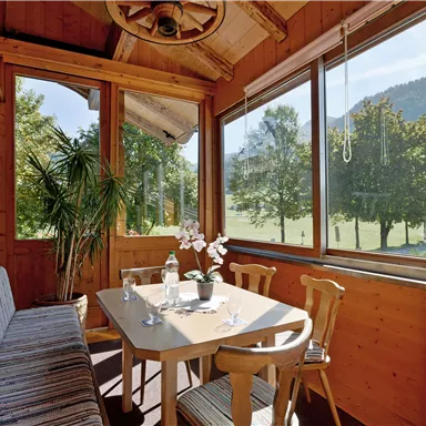 An inviting conservatory with wooden cladding and a stylish table. Large windows provide a view of the green landscape and trees.