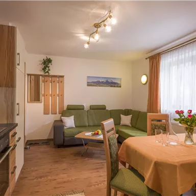 A cozy apartment with a kitchen, a dining area, and a living room. The bright curtains and the flowers on the table give the room an inviting atmosphere.