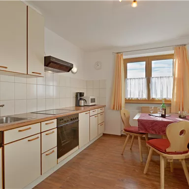 A modern kitchen with white cabinets and wooden details. A dining table with red chairs is next to a window with curtains.