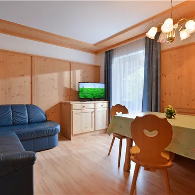 Ein gemütliches Wohnzimmer mit einer blauen Couch und einem Tisch. An der Wand befindet sich ein Fernseher und große Fenster lassen viel Licht herein.