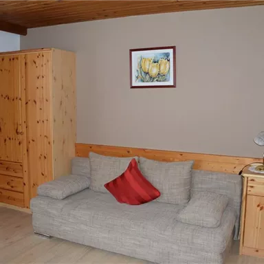 A living room with a bright sofa and a red cushion. In the room, there is a wooden cabinet, a table with a lamp, and a television.