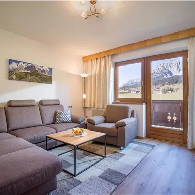 A cozy living room with a modern sofa and an armchair. Large windows offer a view of the mountains and plenty of natural light.