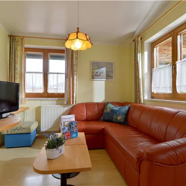A cozy living room with a large red sofa and a table. Large windows let in plenty of light and a television is also present.