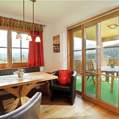 A cozy dining area with a wooden table and a dark bench. Large windows offer a view of the terrace and the surrounding nature.
