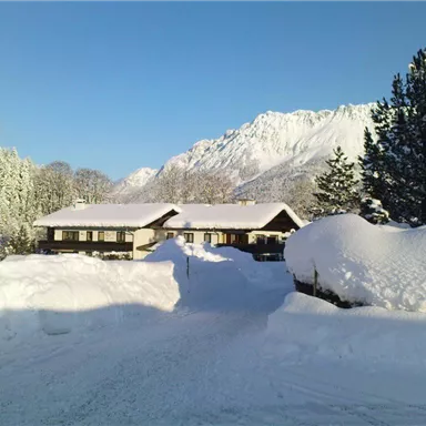 A snow-covered landscape with a cozy house and snow-covered trees. In the background, majestic mountains can be seen under a clear blue sky.