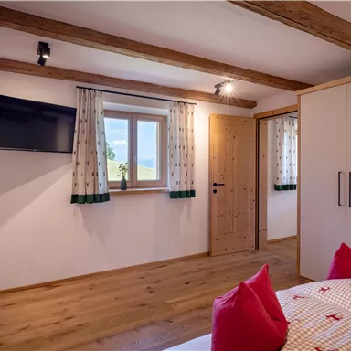 A cozy bedroom with a wooden ceiling and modern amenities. The room is bright and friendly with a large window front and decorative pillows.