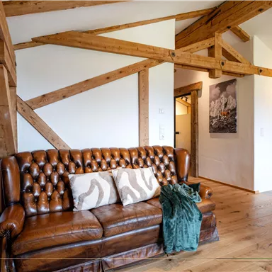 A cozy living room with a brown leather sofa and wooden beams. There is a warm, inviting impression created by the decor.
