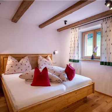 A cozy bedroom with a wooden bed and colorful pillows. Large windows let in plenty of daylight and offer a view of greenery.
