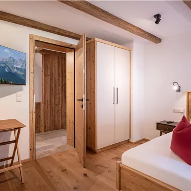 A cozy bedroom with wooden details and a large window. A television hangs on the wall and a table is beside the bed.