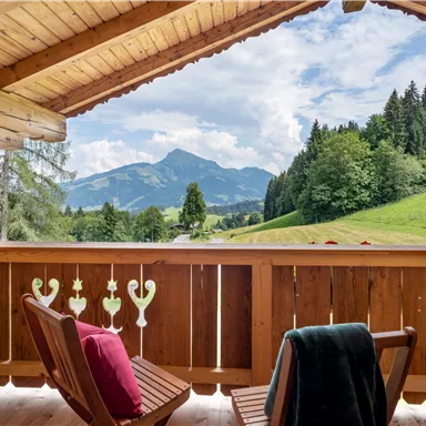 A beautiful view from a wooden balcony of the mountains and the green landscape. Two chairs invite you to relax.