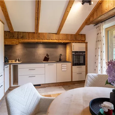 A modern kitchen with wooden elements and white cabinets. The dining area is bright and inviting, with large windows and a round table.