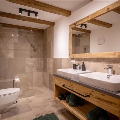 A modern bathroom with wood details, two sinks, and a large mirror. The shower is elegant, and the floor is designed with neutral tiles.