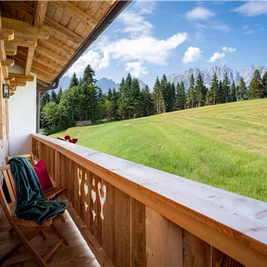 A cozy balcony with a wooden railing and chairs. The view overlooks a green meadow and forested hills under a blue sky.