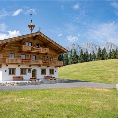A traditional wooden house with several balconies and a beautiful view. In the background, mountains and a blue sky are visible.