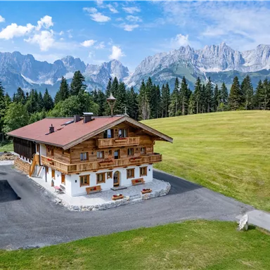 A traditional wooden house surrounded by green meadows and tall mountains. The landscape is picturesque and peaceful.