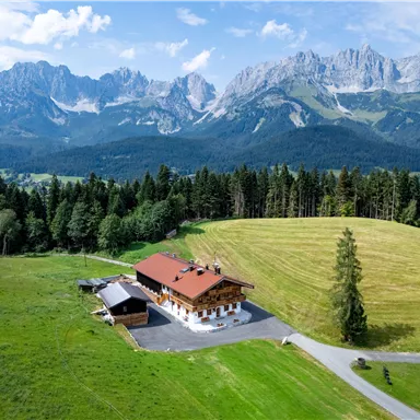 A picturesque mountain cabin surrounded by green meadows and majestic mountains. The sky is clear and bright blue.