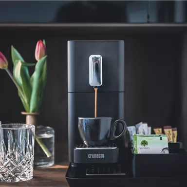 Eine moderne Kaffeemaschine steht auf einem Tisch neben zwei Gläsern und einer Vase mit Tulpen. Daneben befindet sich ein Tablett mit Teebeuteln und Zucker.