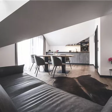 Ein modernes, helles Wohnzimmer mit einer stilvollen Couch und einem Esstisch. Der Raum hat große Fenster, die für viel Tageslicht sorgen.