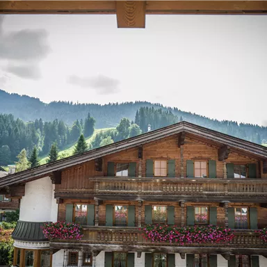 A traditional wooden house with colorful flower balconies and a view of the mountains. The sky is cloudy and the surroundings are green and hilly.
