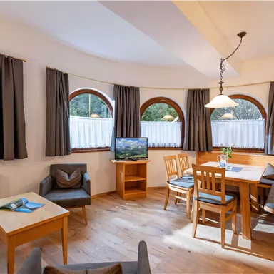Ein gemütlicher Wohnbereich mit Rundfenstern und Holzmöbeln. Es gibt einen Tisch mit Stühlen und eine Sitzecke, sowie einen Fernseher.