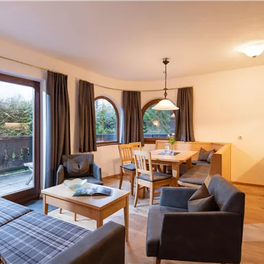 Ein gemütlicher Wohnbereich mit einem Esstisch, Stühlen und einer Couch. Große Fenster sorgen für viel Licht und einen schönen Blick ins Freie.
