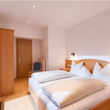 Ein modernes Schlafzimmer mit einem Doppelbett und zwei Kissen. Es gibt einen Holzschrank und einen Stuhl in einem klaren, minimalistischen Design.