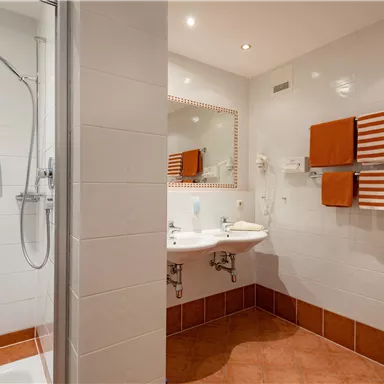Ein modernes Badezimmer mit einer Dusche und zwei Waschbecken. Orangefarbene Handtücher und eine hellgraue Fliesenwand schaffen eine angenehme Atmosphäre.