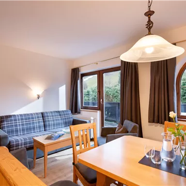 Ein gemütliches Wohnzimmer mit einem Sofa, einem Tisch und Stühlen. Durch das große Fenster fällt viel Tageslicht und bietet einen Blick ins Grüne.