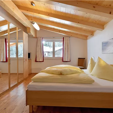 Ein gemütliches Schlafzimmer mit Holzböden und einer Holzdecke. Das Bett ist mit gelben Kissen und Decken dekoriert, und es gibt große Fenster mit einem Blick nach draußen.