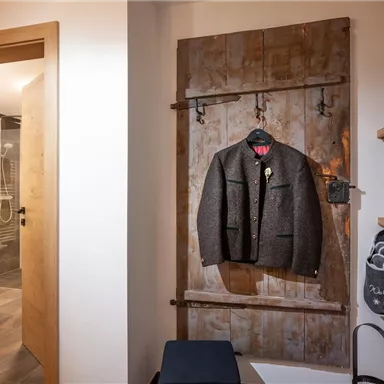 Ein eleganter Anzug hängt an einer rustikalen Garderobe in einem modernen Raum. Im Hintergrund ist ein Badezimmer mit einer Dusche sichtbar.