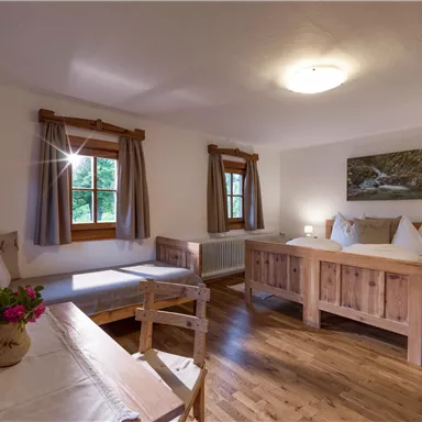 Ein gemütliches Zimmer mit zwei Holzbettgestellen und einem Tisch. Helle Vorhänge und ein großes Fenster sorgen für eine einladende Atmosphäre.