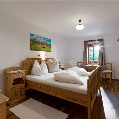 Ein gemütliches Schlafzimmer mit einem Doppelbett aus Holz und vier Kopfkissen. Große Fenster sorgen für Tageslicht und Blick auf die Natur.