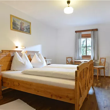 Ein gemütliches Schlafzimmer mit einem Holzbett und zwei Kopfkissen. Dazu gibt es eine Kommode, einen kleinen Tisch und Stühle, sowie ein Fenster mit Gardinen.