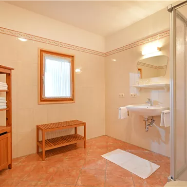 Ein helles Badezimmer mit Fliesenboden und Dusche. Ein Holzregal mit Handtüchern und ein Waschbecken sind ebenfalls zu sehen.