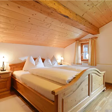 Ein gemütliches Schlafzimmer mit Holzmöbeln und einem geneigten Dach. Das Zimmer ist hell und freundlich mit Fenster und Vorhängen.