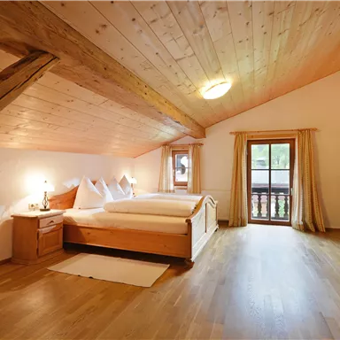 Ein gemütliches Schlafzimmer unter dem Dach mit Holzdachbalken. Das Zimmer verfügt über ein Doppelbett, Holzfußboden und einen Balkon mit Aussicht.