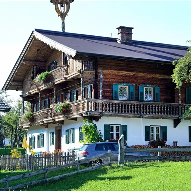 Ein traditionelles Holzhaus mit einem schönen Balkon und bunten Fensterläden. Es steht auf einer Wiese mit grünem Gras im Hintergrund.
