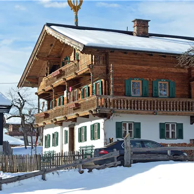 Ein traditionelles Holzhaus im Schnee mit grün gestrichenen Fenstern und einem schönen Balkon. Der Himmel ist blau und die Umgebung ist winterlich.