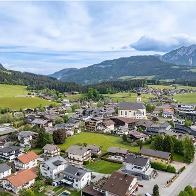 Ein malerisches Dorf in den Bergen mit traditionellen Häusern und grünen Wiesen. Im Hintergrund sind majestätische Berge und ein klarer Himmel zu sehen.